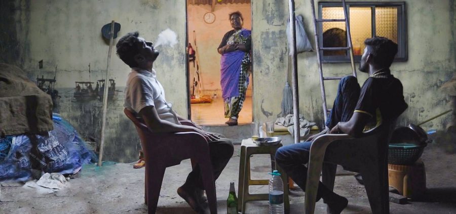 Two Koli men sit outside a house on plastic chairs sharing a meal and beers. Between them an older Koli woman wearing a purple and gold sari stands in the doorway of the home, her hands crossed around her stomach and a smile on her lips as she is lit by the warm golden light inside. One of the men, wearing a collared shirt and trousers, blows cigarette smoke from his mouth as he looks up into the sky. The other man, wearing a black soccer shirt and blue pants, turns away from the camera as he looks towards the woman in the doorway.