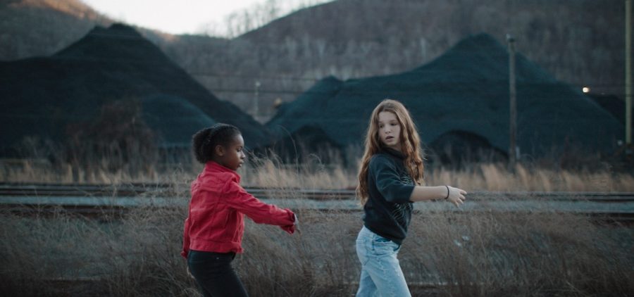 A young white girl and a young Black girl raise their arms in a coordinated dance beside an empty railroad track. On the opposite side of the track, mounds of coal and the mountains of Appalachia fill the landscape.