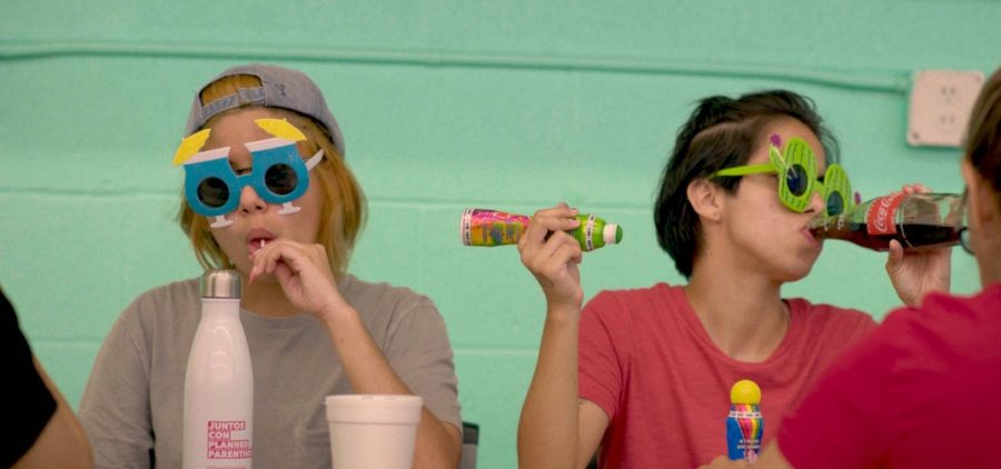 In a vibrant setting, two Latinx teens enjoy a light-hearted moment. One, donning a cap and novelty piña colada sunglasses, sucks on a lollipop, while the other, wearing a red tee and novelty cactus glasses, sips from a soda bottle. The aqua wall behind them adds to the playful atmosphere.
