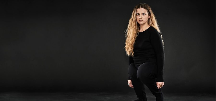 A white woman with long wavy blonde hair and unusually short legs wears an all black outfit and stands against a black backdrop. She looks into the camera with a confident gaze.