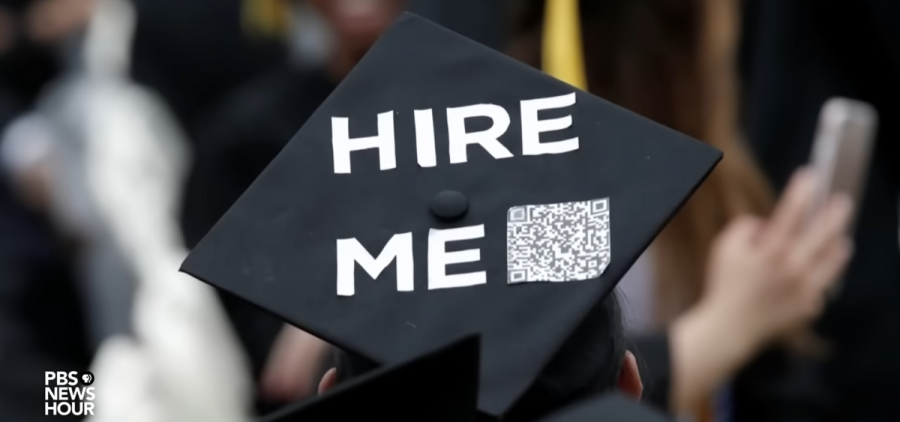 A student wears a graduation cap that reads "HIRE ME" and has a QR code.