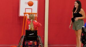 A girl in a wheelchair shooting a basketball into a hoop with the help of a volunteer at the SOCIL Adaptive Sports Palooza.