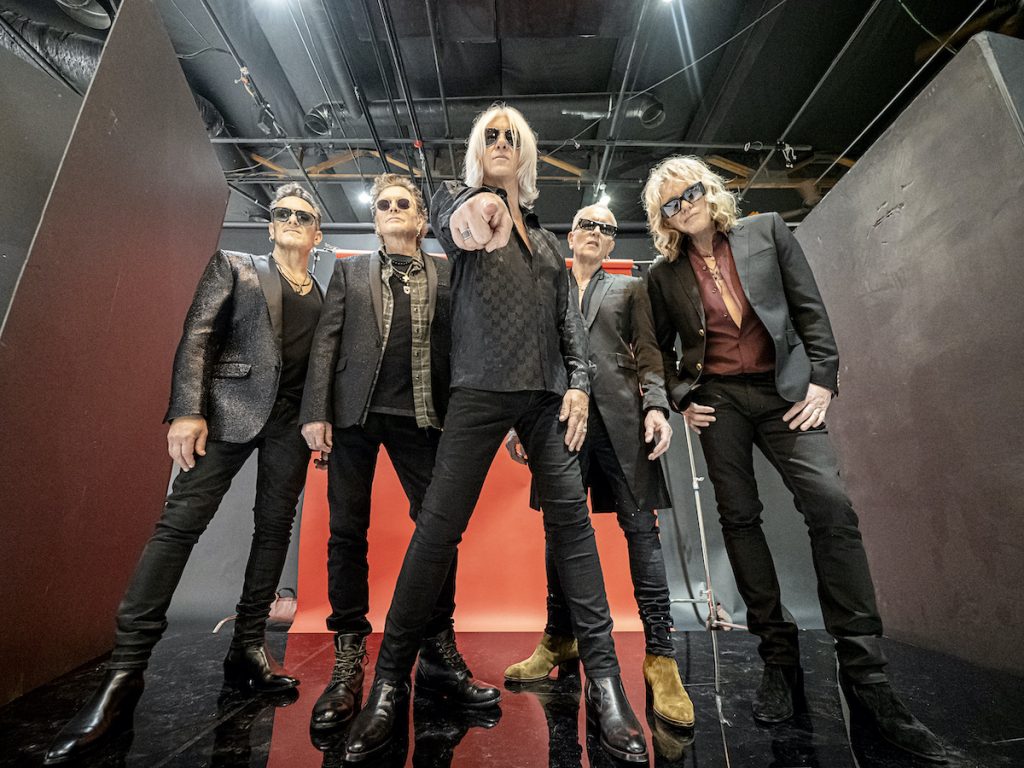 A promotional photo of the band Def Leppard. It is taken from a vantage point below the band, and they are pointing at the camera. All of the four members have blonde hair and are fair skinned and slim. 