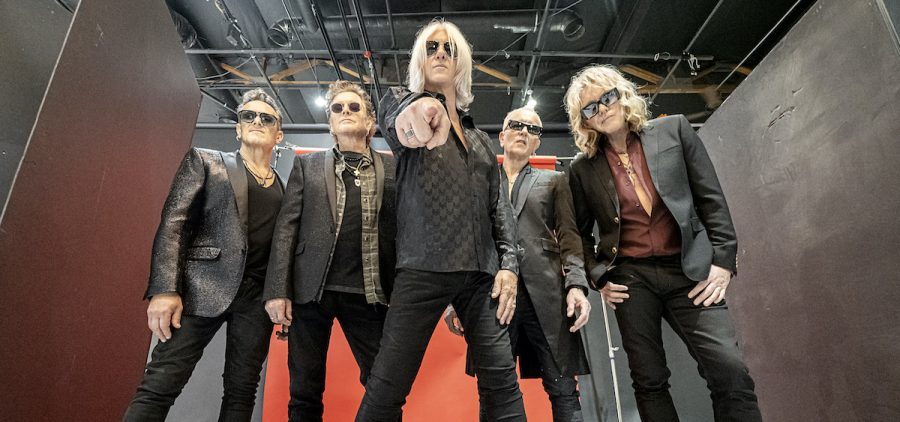 A promotional photo of the band Def Leppard. It is taken from a vantage point below the band, and they are pointing at the camera. All of the four members have blonde hair and are fair skinned and slim.