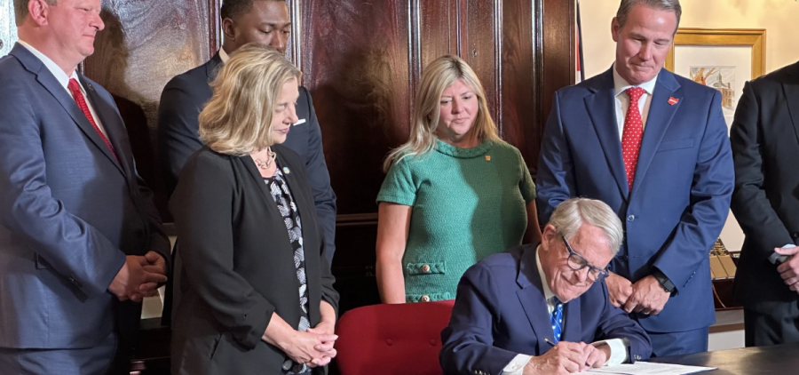 Ohio Governor Mike DeWine signs a bill into law while other people stand behind him.