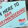 A man in a hat holds a large teal sign reading "SIGN HERE TO BAN GERRYMANDERING."