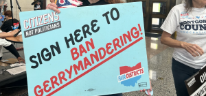 A man in a hat holds a large teal sign reading "SIGN HERE TO BAN GERRYMANDERING."