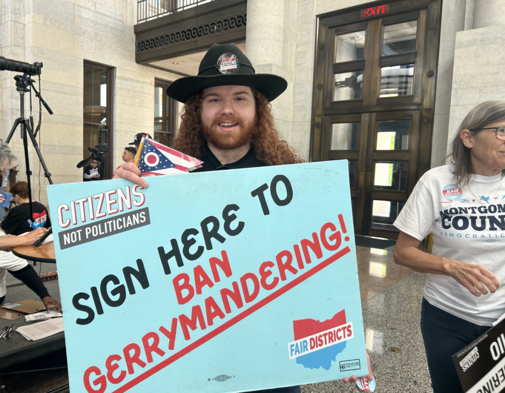 A man in a hat holds a large teal sign reading "SIGN HERE TO BAN GERRYMANDERING."
