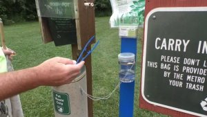 Next to a trash bag station for your pets is a tick collection station at a park. A clear jar of alcohol is attached to a pole and full of ticks. A pair of tweezers dangles from the post for parkgoers to pluck ticks off of themselves.