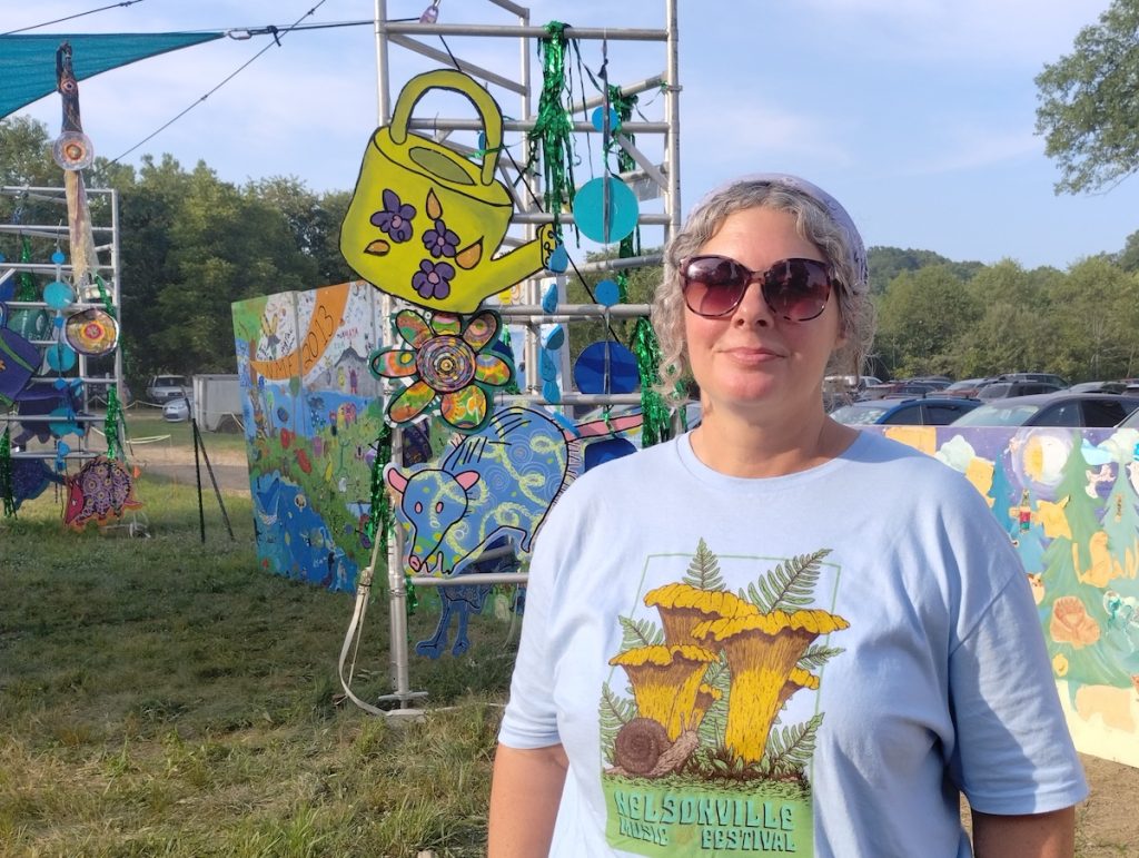 Desirae Matherly. A woman wearing a blue shirt is standing outside with colorful art advertising the Nelsonville Music Festival. 
