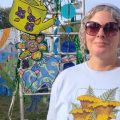 Desirae Matherly. A woman wearing a blue shirt is standing outside with colorful art advertising the Nelsonville Music Festival.