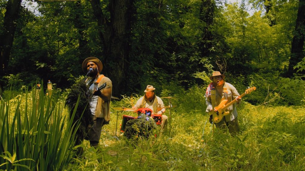 A screen grab from the music video for an indie rock song. Three musicians are playing their instruments outside in the wilderness. 