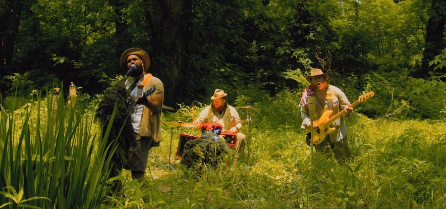 A screen grab from the music video for an indie rock song. Three musicians are playing their instruments outside in the wilderness.