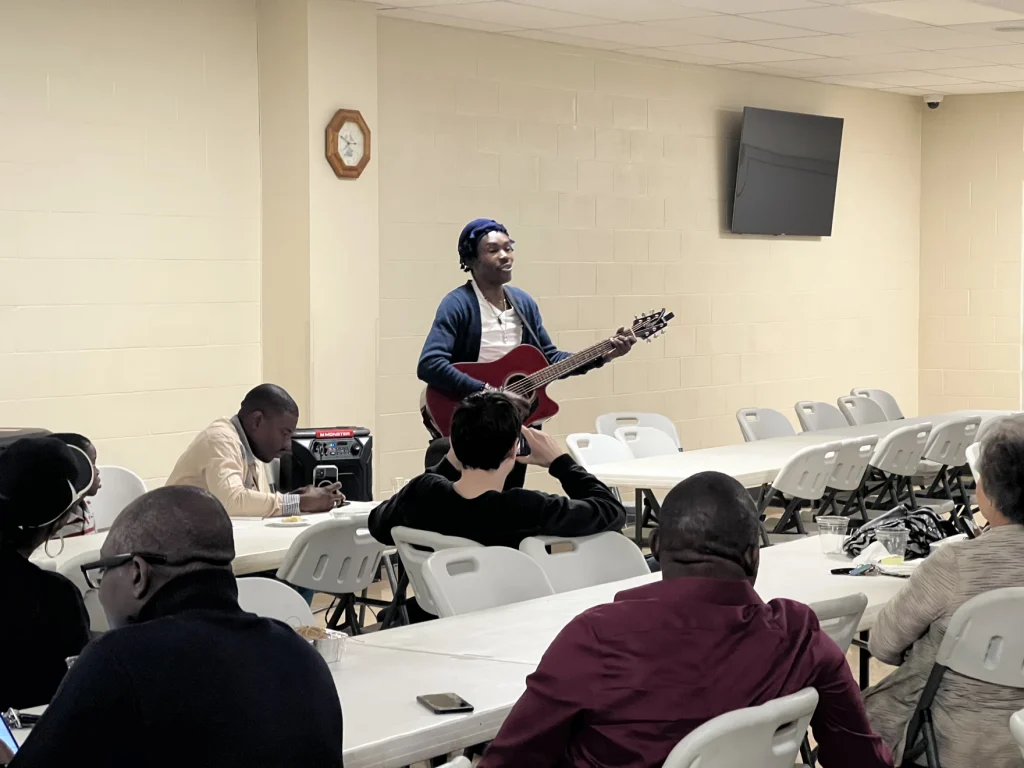 Ams Marcelin sings and plays guitar in front of a crowd at the community center.