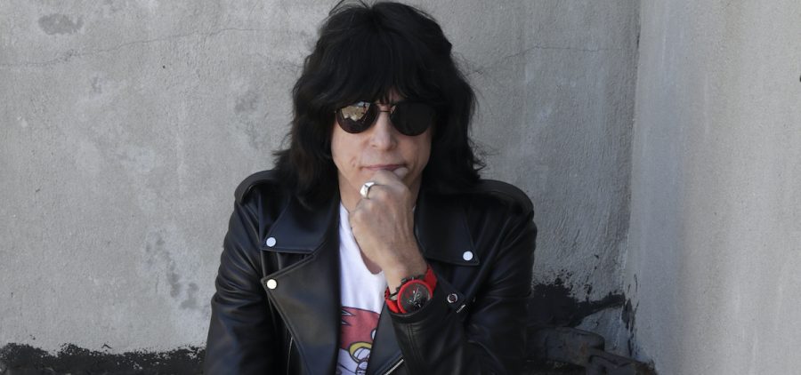 A press photo of Marky Ramone. He is seated on a backwards chair. he has black hair and sunglasses, and a motorcycle jacket.