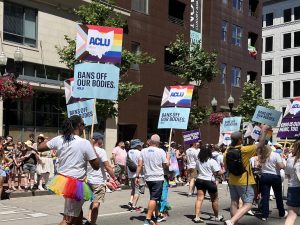 A crowd of demonstrators march down a street holding signs with ACLU printed over a Pride flag and the words "Bans off our bodies."