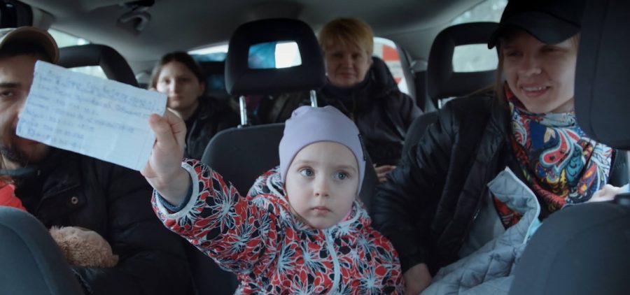 Inside a crowded vehicle with a blurred window view, a white child in lavender beanie and colorful sweater looks to the side with plaintive expression holding a slip of paper. Surrounding the child are white adult passengers, some of whom look to the child and others to the camera.