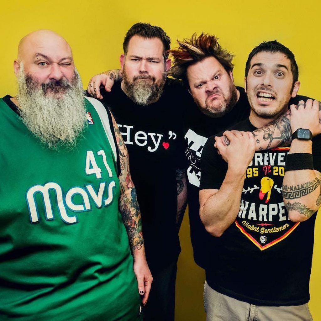 Four men pose against a plain yellow background. 