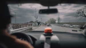 Through the front windshield of a car the blurred silhouette of a driver with a large orange siren in the middle of his dashboard looks out onto a grey, foreboding sky, and a highway. A few feet up the highway is a roadblock. The roadblock looks hastily thrown together with wooden panels and bags of gravel. More bags litter the side of the road and two flags fly above the roadblock, their colors not visible.