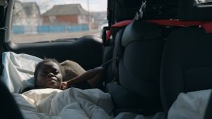 A young Black woman lies across several seats in the back of a van. She has a white pillow to support her head and a white comforter over her body. She looks up at the camera with worry as she leans her elbow against the drivers seat. Above her, the car's window looks out onto houses and a blue sky, slightly out of focus.