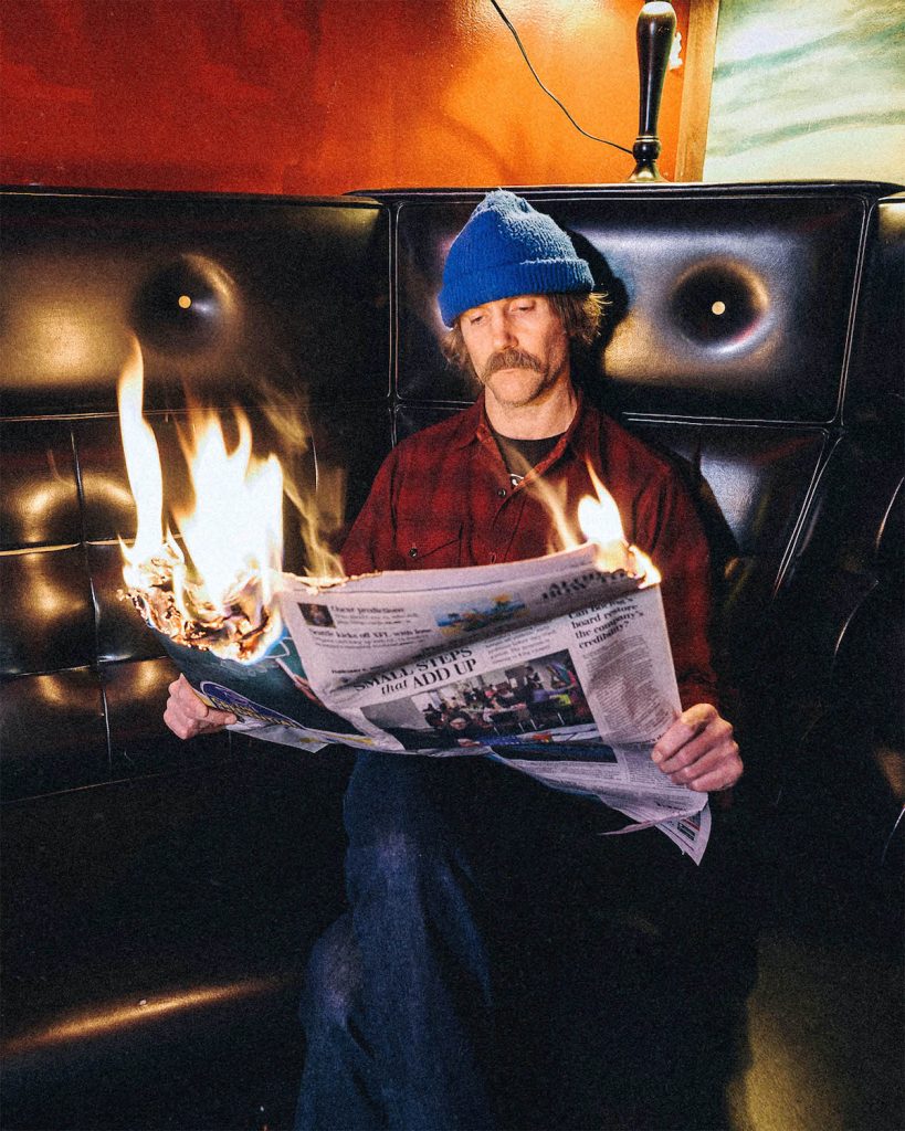 A promotional photo of Dean Johnson. He is sitting down and reading a newspaper that is on fire and he is wearing a blue cap. 