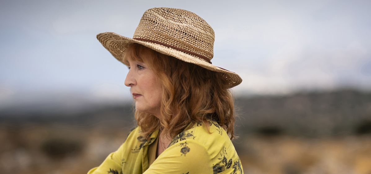 Susan Ryeland (Lesley Manville) sitting on beach with yellow shirt and straw woven hat Photographer: Jonathan Hession Courtesy of Eleventh Hour Films and MASTERPIECE.