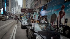 In a bustling Hong Kong city street, a campaign rally takes place. A few Hong Konger individuals stand through the sunroofs of cars, one holding up a sign with a portrait of a candidate and a number 8. The scene is set against a backdrop of tall buildings, with a city bus to the right displaying a large advertisement.