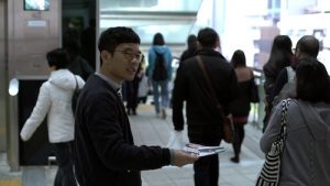 A Hong Konger man with short hair and glasses hands out campaign flyers at at the entrance of subway station in a sunny city. Commuters walk past him, their backs to the camera, none seeming to meet his gaze. The man wears an eager smile on his face.