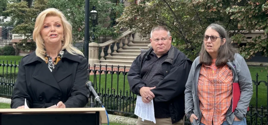 A woman speaks at a podium with a man and a woman behind her to the right.