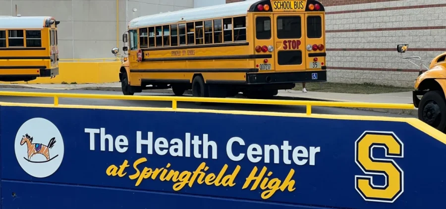 Busses pull up to the front of a school with a sign in front of them highlighting the Health Center at Springfield High