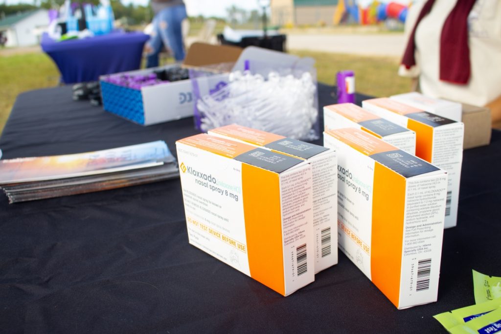 A table at a fairground is set up with boxes of Naloxone and other giveaways.