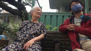 An elderly woman sits on a bench with a young man in an open red jacket, wearing a handmade blue fabric face-mask and looking at the distance, above the frame, slightly content. The woman speaks to him. Details of the playground are visible in the background: a blue structure, greenery.