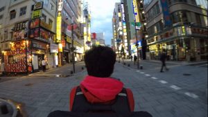 A third person omniscient wide angle shot of a man riding a bicycle wearing a big, thermal Uber Eats delivery backpack, camera affixed to the bag. Tokyo streets engulf him left, right and above, lights blurred by the motion, yet suggesting a myriad open restaurants and stores, few passersby.