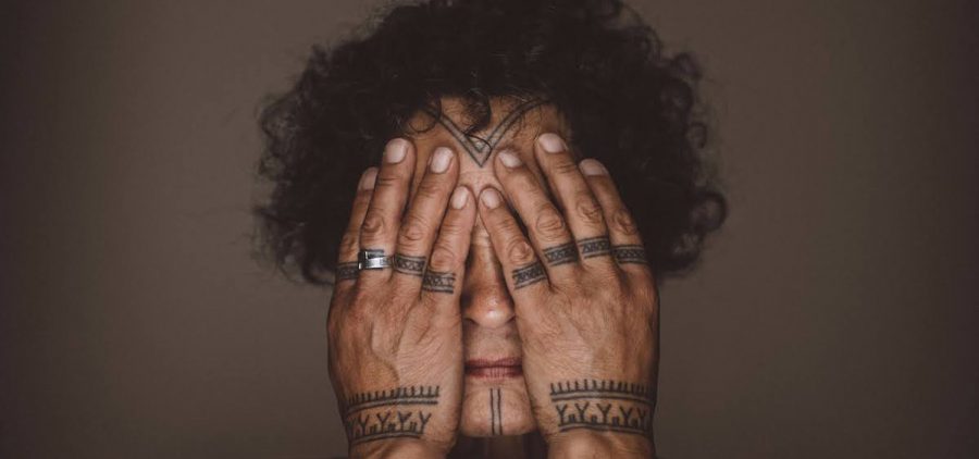 An Inuit woman with curly dark hair and a tattooed chin and forehead conceals her eyes behind tattoo-adorned hands against a brown background.