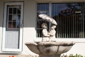 A bird bath with two stone children under an umbrella sits outside a building with an OPEN sign in the window.