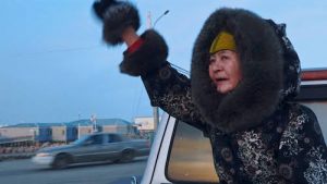 An Inuit woman with a tattooed chin raises her a mittened hand to wave to someone. She wears a black jacket with a white floral pattern, fur lined hood and cuffs, and a yellow hat. The hood is up on her head surrounding her face with dark fur. She stands behind a silver car and in the background another car drives by grey houses and a cloudless blue sky.