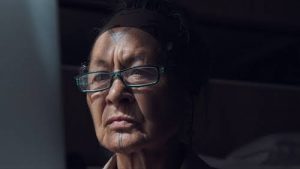 An Inuit woman with tattooed chin and forehead looks into the distance with a stern expression. She wears blue reading glasses and a black headband. Behind her dark shadows reach out to somewhat obscure her face.