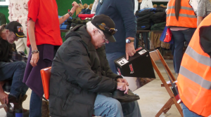 A veteran, wearing a newly acquired coat, puts his shoes back on after trying on new boots.