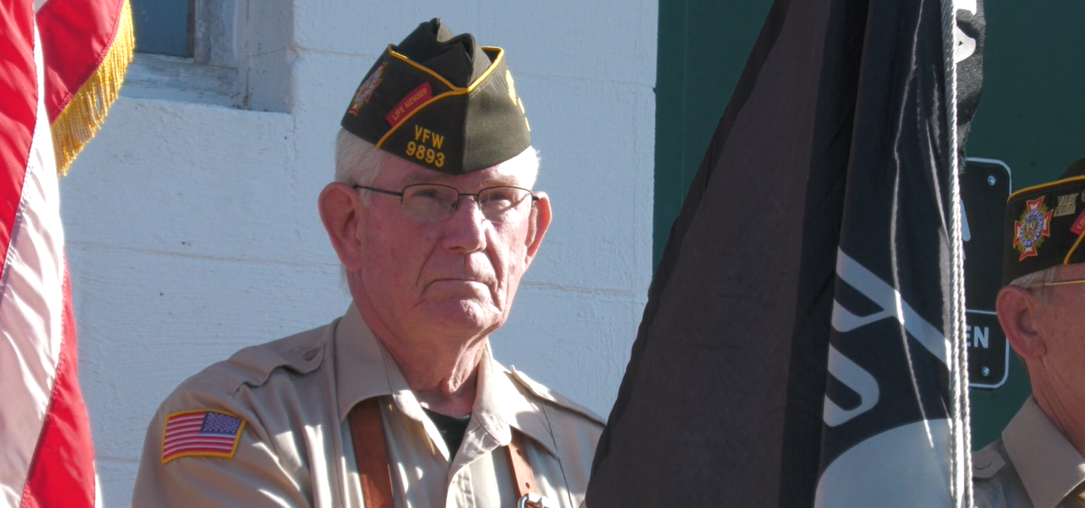 A veteran holds a POW flag for the Athens Area Stand Down opening ceremony. He stands between an American flag and another veteran.