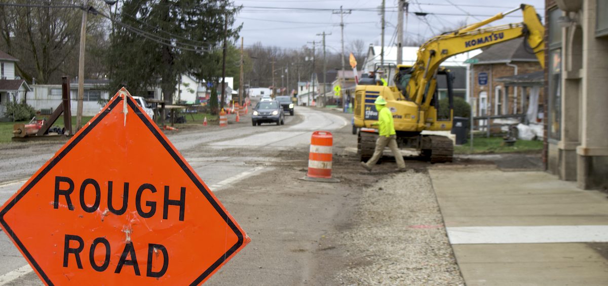 A backhoe operator digs up a street in Chauncey so the sewer line underground can be replaced. [David Forster | WOUB]