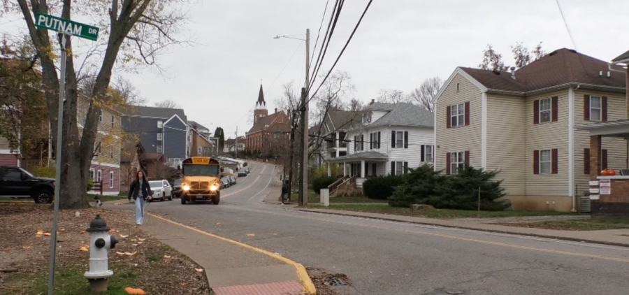 Mill Street homes line the road as a bus comes through and people walk on the sidewalk.