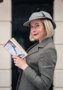 Lucy Worsley in deerstalker hat and book in hand in front of the 221B Baker Street door