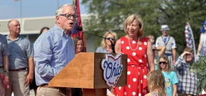 Gov. Mike DeWine at the Ohio State Fair speaks at a podium with his wife in the background smiling