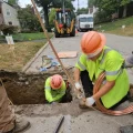 A crew of men in orange hard hats and yellow jackets work in a hole carved out of a street.