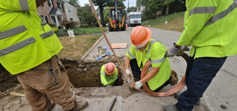 A crew of men in orange hard hats and yellow jackets work in a hole carved out of a street.