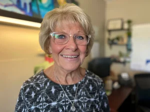 An older woman with blond hair and glasses smiles for a portrait