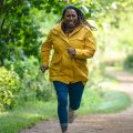 Suzie Harris (Jo Martin) from MASTERPIECE The Marlow Murder Club running down dirt path in jeans and yellow rain coat looking distresses