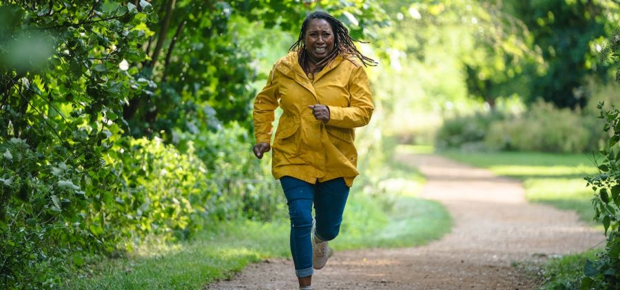 Suzie Harris (Jo Martin) from MASTERPIECE The Marlow Murder Club running down dirt path in jeans and yellow rain coat looking distresses