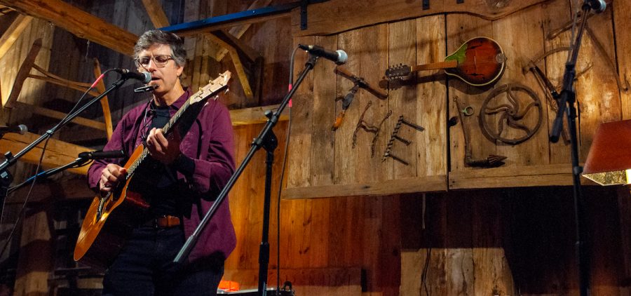 A man plays guitar and sings into a microphone on a stage in a barn.