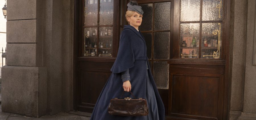 Shown: Eliza Scarlet (Kate Phillips) Photographer: Sever Zolak Detective Miss Scarlet in dark blue cloak and gray cap holding brown leather bag in front of wood covered store front